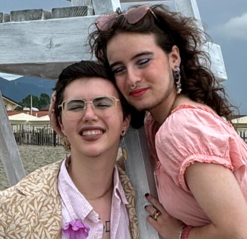 Dmitri and Reuven smiling on a windy beach day in Massa on the Ligurian Sea coast in northern Italy. Dmitri is wearing a tan olive-patterned blazer and pink dress shirt, Reuven is wearing a pink dress and moon+star earrings from lovely Phyllis.