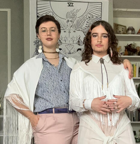 Art Museum Eleganza 2024 Dmitri in a white fringe shawl, blue linen dress shirt and dusty pink slacks, sporting handmade antique watch clock-face earrings, and Reuven in a white fringe western shirt with a cicada bolo tie and a dusty pink western skirt from Dmitri's Grammy Eileen.
